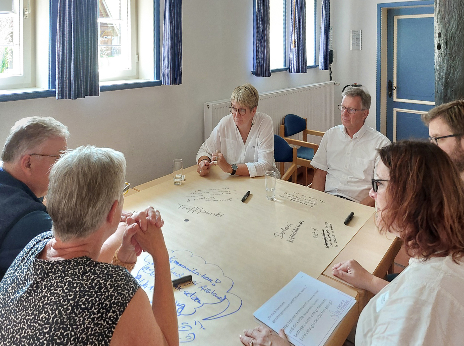 Sechs Personen sitzen am Tisch, reden miteinander und machen sich Notizen.